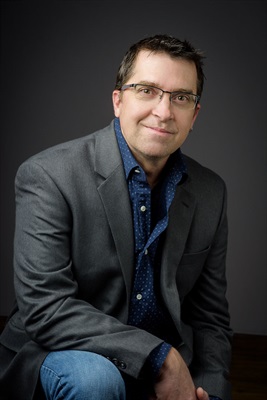white male with brown hair and glasses dressed in jeans, a dark button down shirt and a grey blazer