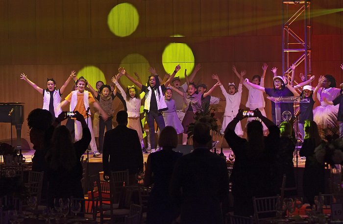 Dozens of Disney Musicals in Schools students stand with hands up during a Broadway Ball performance. People in the front are on their feet snapping photos of the kids in costume.