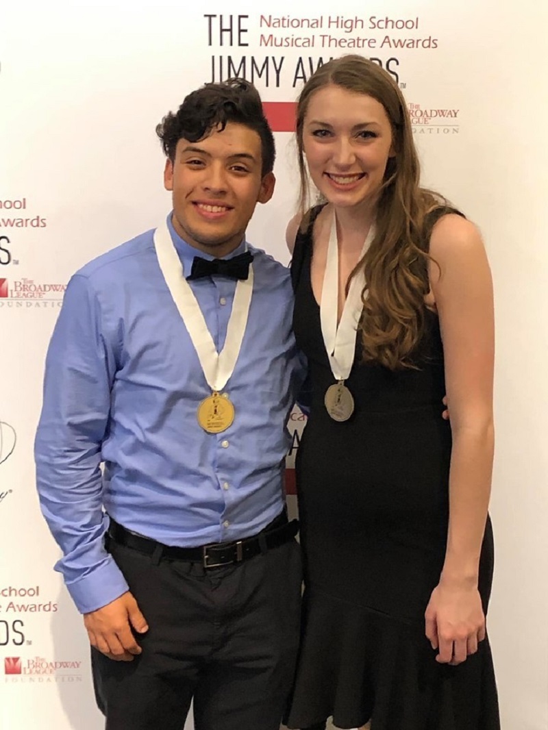 A young man in a blue shirt and a young woman in a black dress stand together for a photo, medals around their necks.