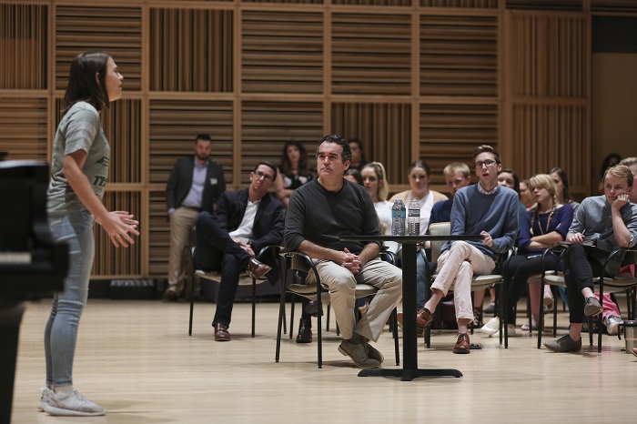 Brian d'Arcy James sits at a small table in the Scott Recital Hall as a student sings a song for the whole room. Students watch from their seats behind Brian. 