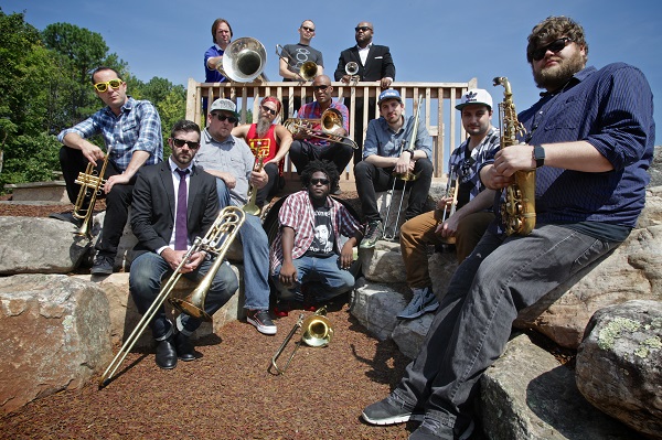 12 laid back members of the No BS! Brass band, some wearing sunglasses and flat-billed caps, sit with their instruments at a sunny park.  