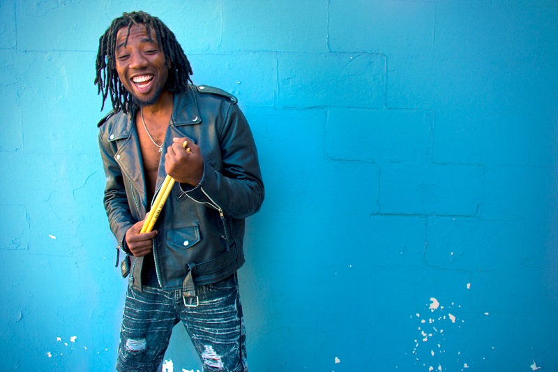 A young African American man stands against a blue wall, smiling with drumsticks in his hands