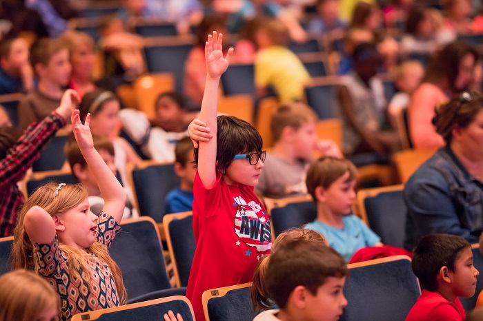 A young child wearing glasses stands up and raises his hand during a show at the Holland Center. Other children sitting around him are also raising their hands.
