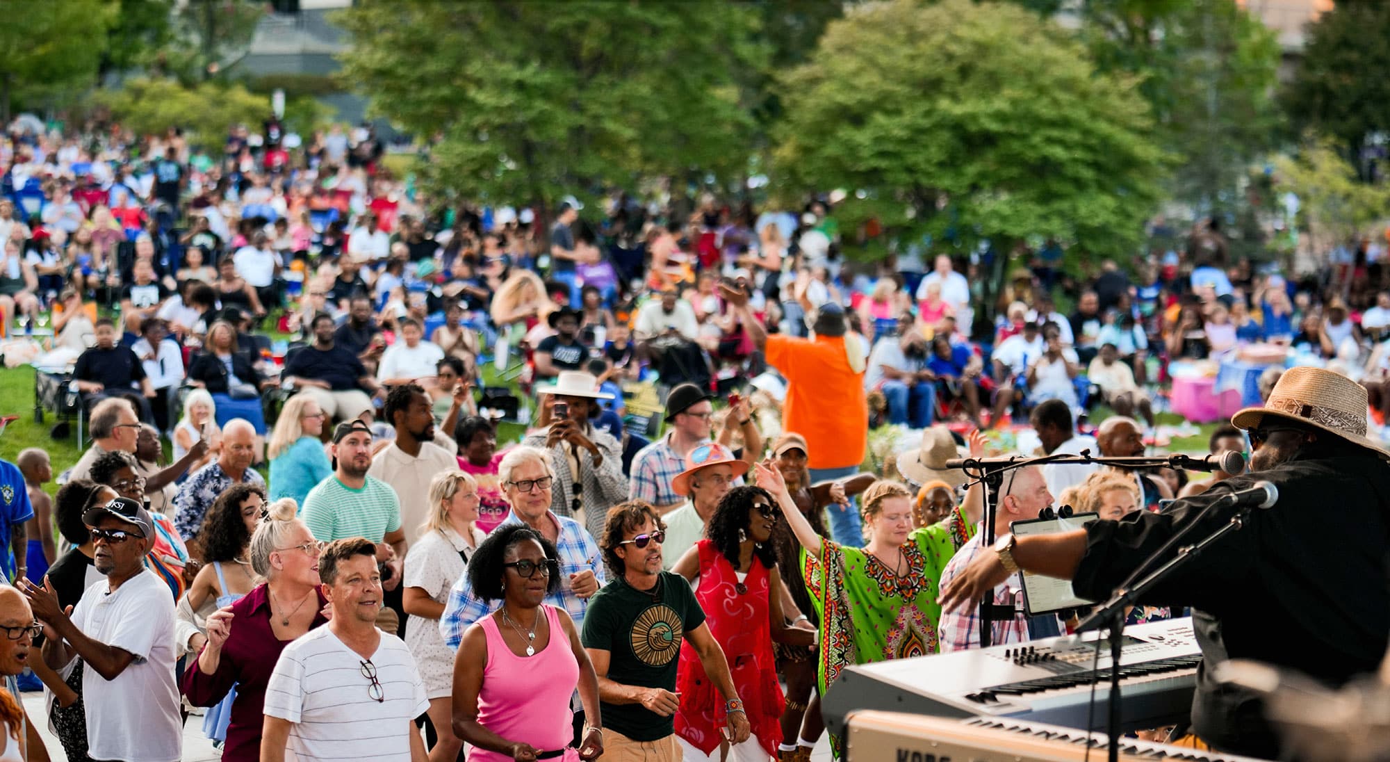 A musician performs on a keyboard and sings into a microphone on an outdoor stage, engaging a lively and diverse crowd of people dancing, clapping, and enjoying the music in a large park filled with attendees seated on the grass and in lawn chairs.