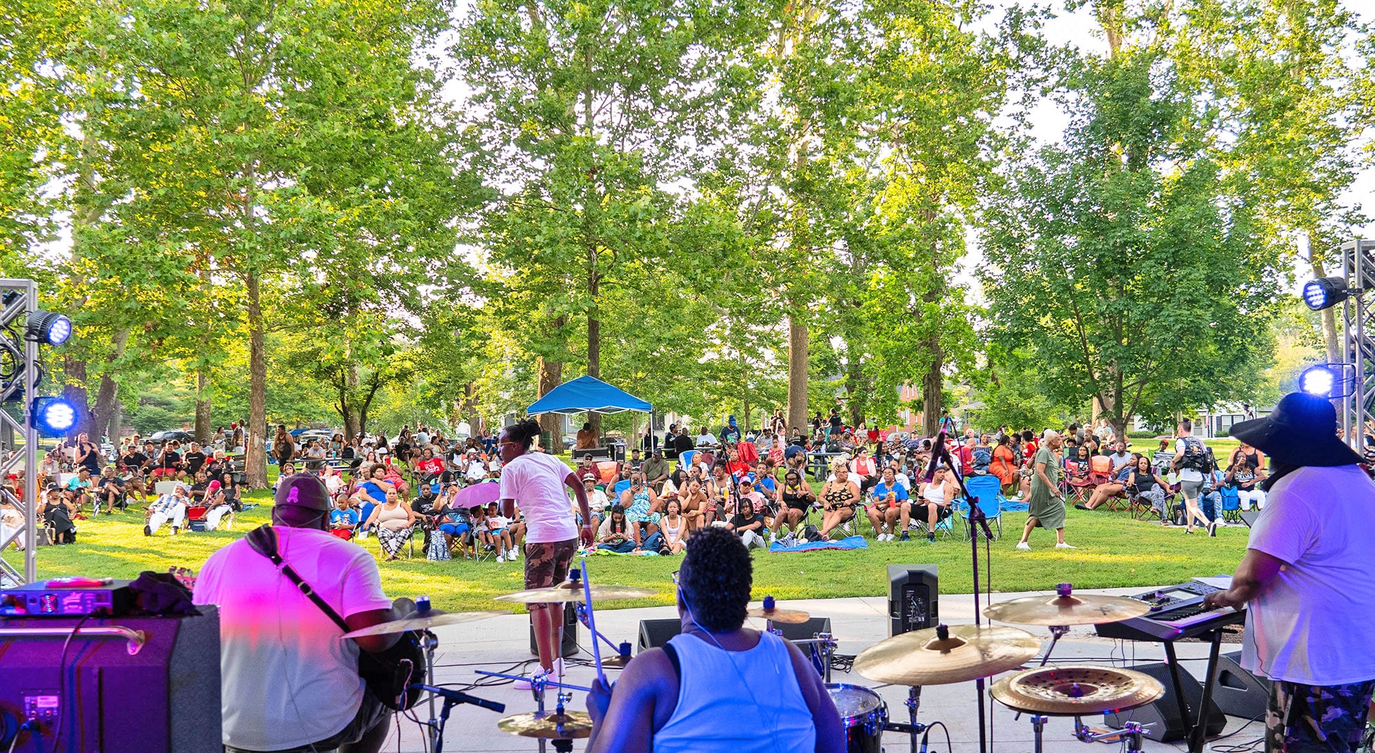 A live band performing on an outdoor stage in a park, with a large, diverse crowd seated on the grass and in lawn chairs under the shade of tall green trees on a sunny day.