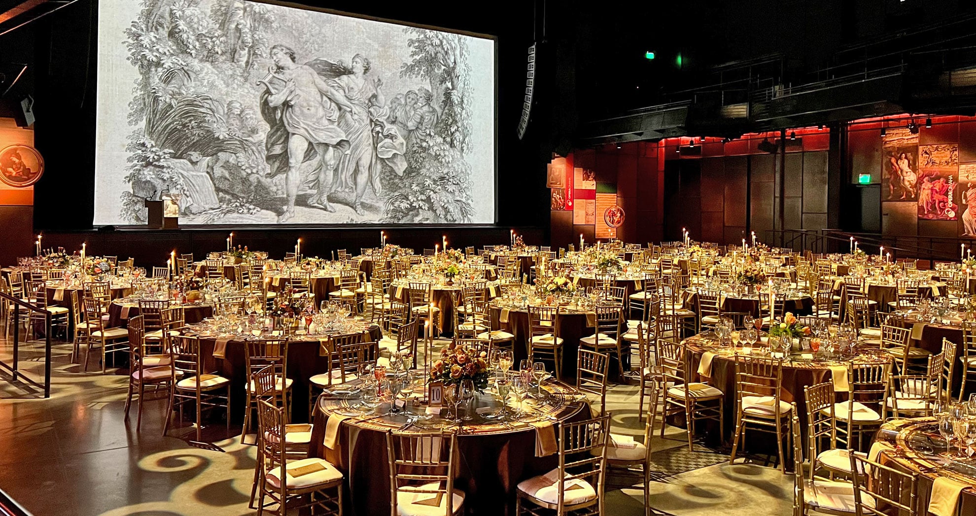 a seated dinner is set up with round tables that fill the room. A large screen fills the back of the room depicting classical black and grey artwork.