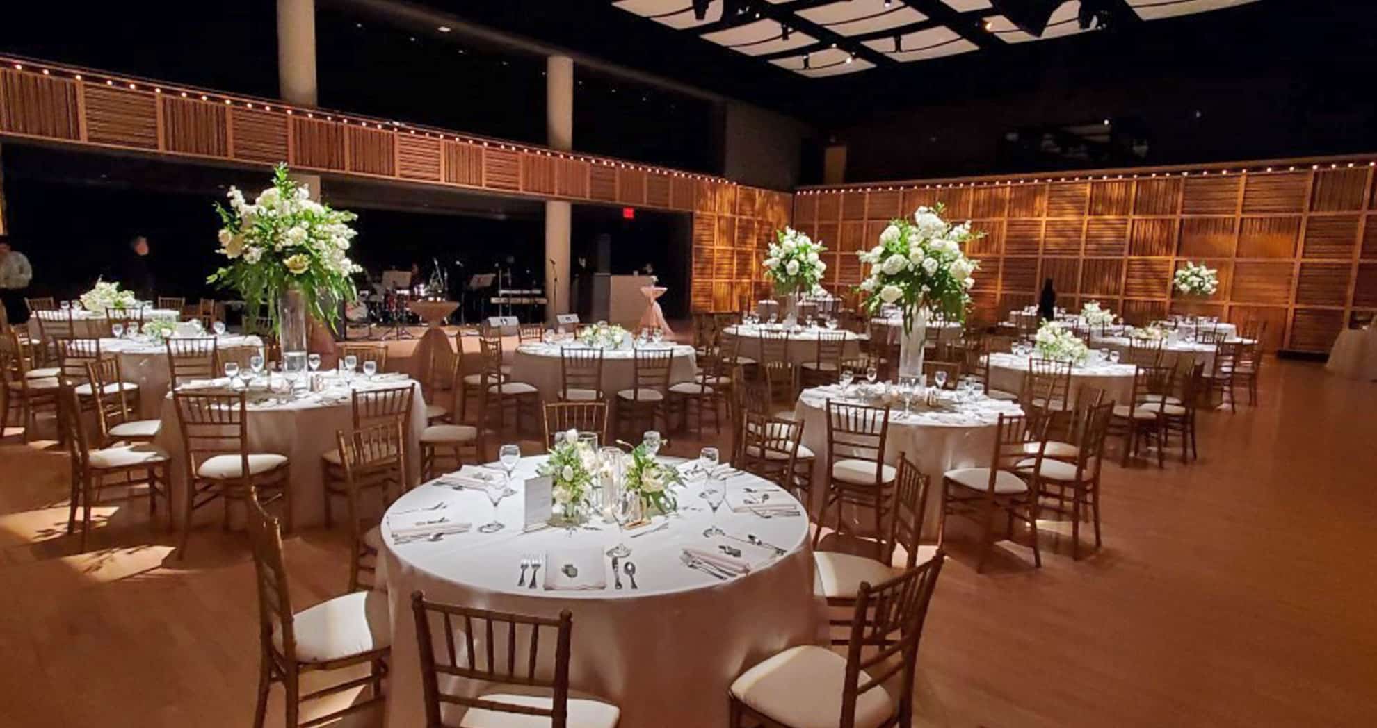 a reception set up in a dimly lit room. round tables are covered in white cloths and have green foliage center pieces