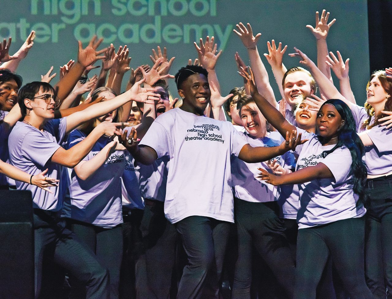 students performing on stage in light purple t shirts, most centered around a young man center stage singing