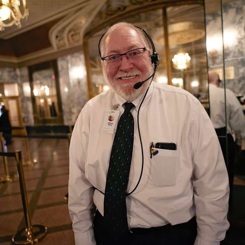 an elderly white man standing in a lobby with a head set on. he wears a white long sleeved shirt and a black tie.