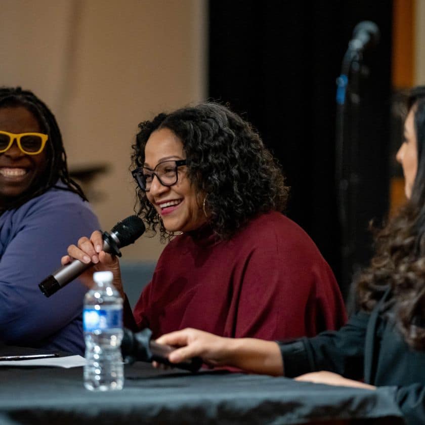 three women of color sit at a desk on a panel, the woman in the center is smiling while she speaks into a microphone