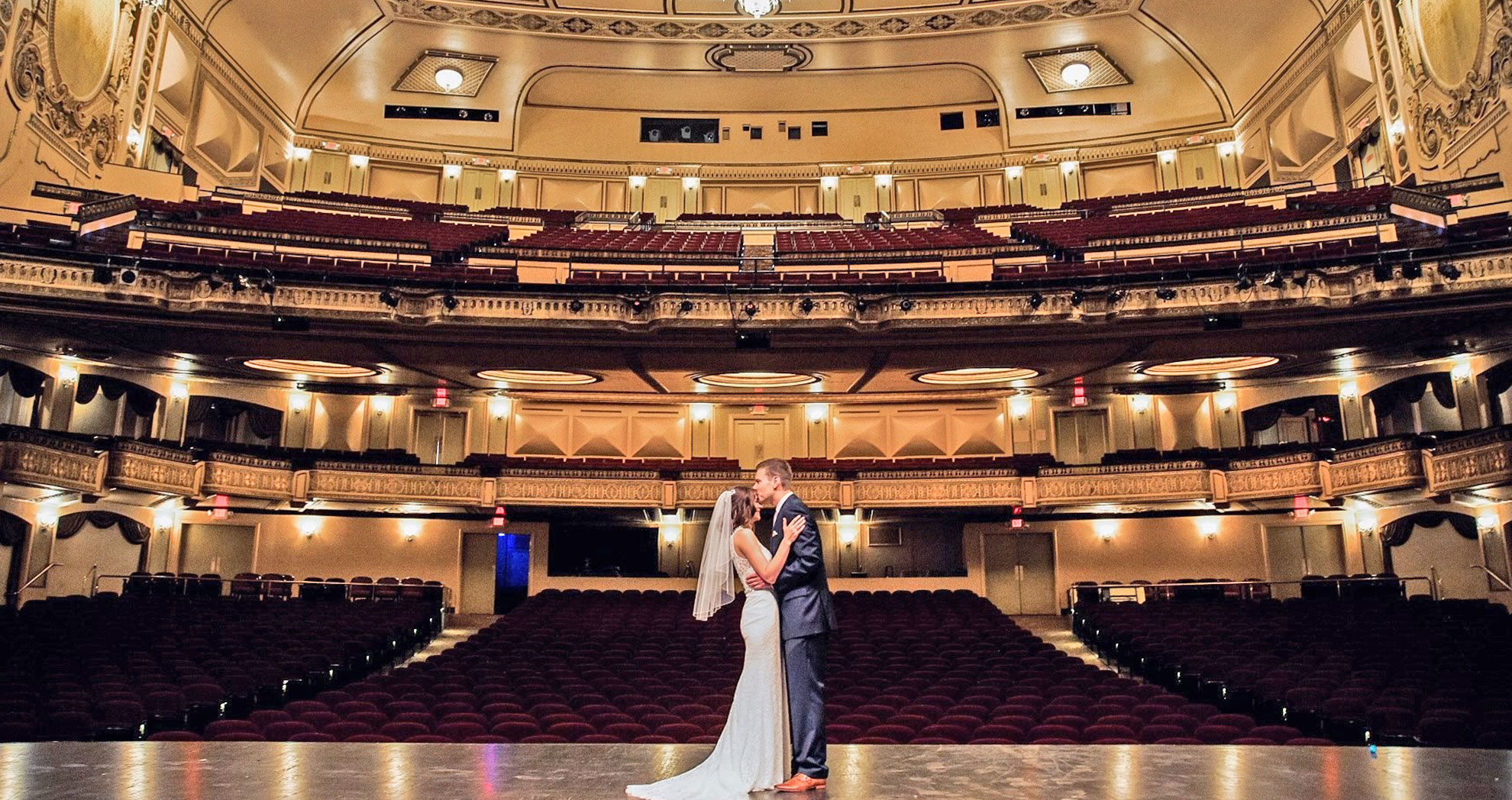 a bride and groom stand center stage in a theater with the house behind them while they embrace