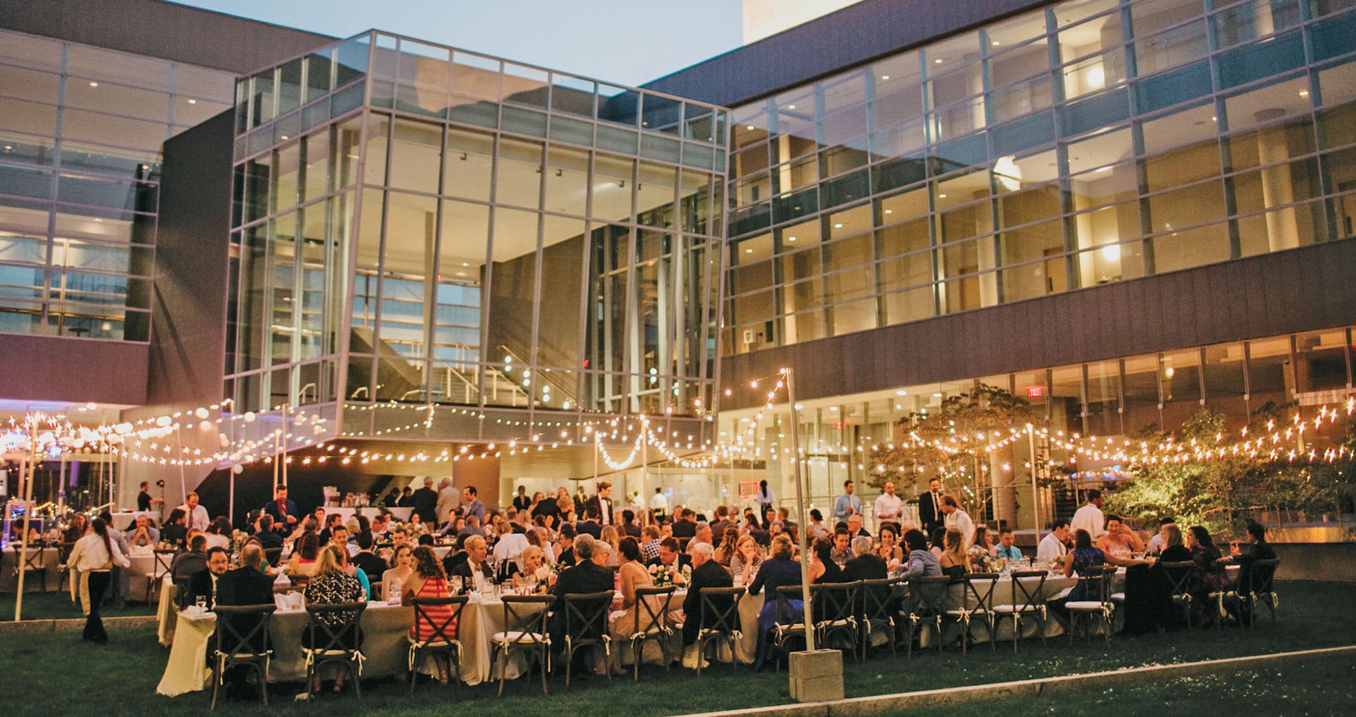 a wedding celebration in a courtyard in the evening with string lights and a crowd seated for dinner