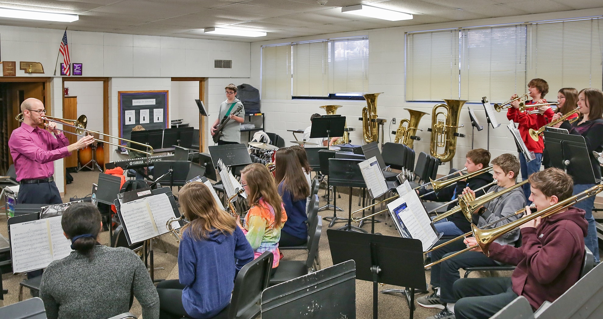 Music teacher in a wine color shirt playing the trombone while teaching a group of students in a school music room. The students, holding various brass and wind instruments, are seated in rows with sheet music on stands in front of them.