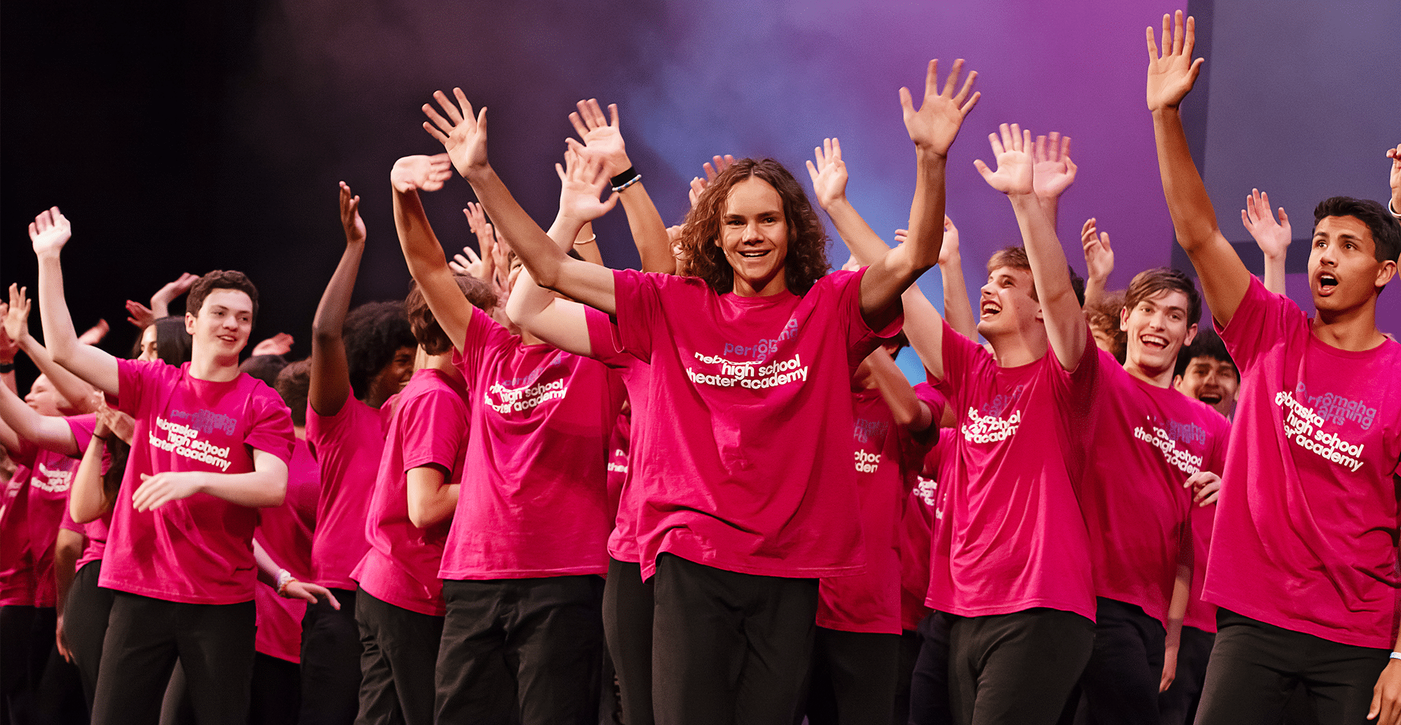Nebraska Theater Academy students on stage wearing pink shirts and waving